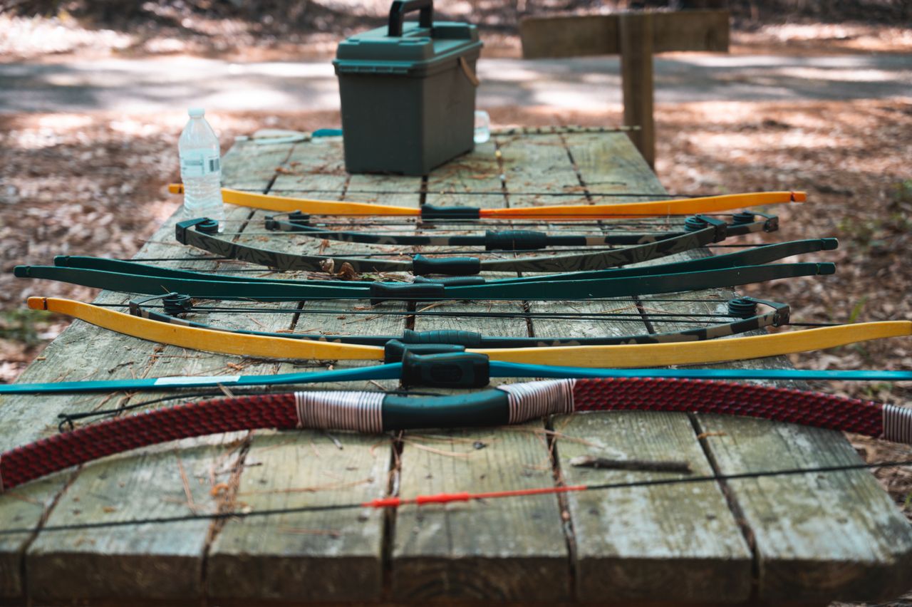 Archery bows and arrows laid out on a wooden table in a forest setting, showcasing vibrant colors.