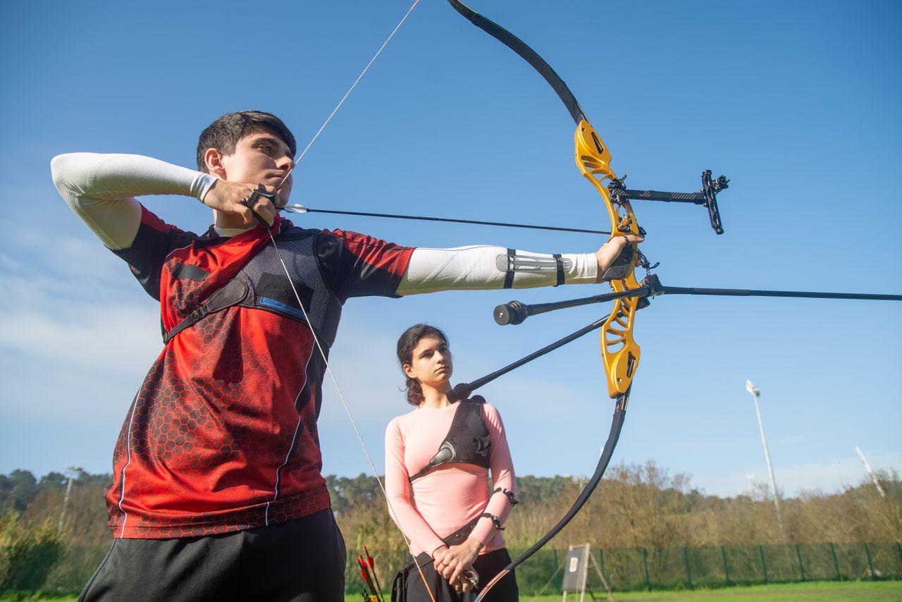 Two young adults practicing archery with a recurve bow in a sunny outdoor setting in Portugal.