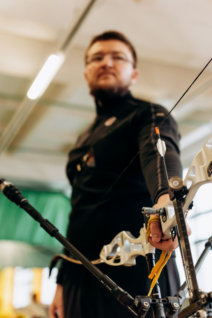 An archer wearing black concentrates indoors while preparing to release an arrow.