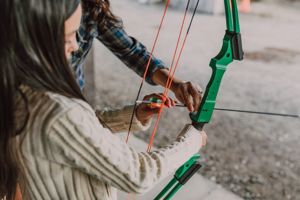 A young girl learning archery with a green recurve bow outdoors, assisted by an adult.