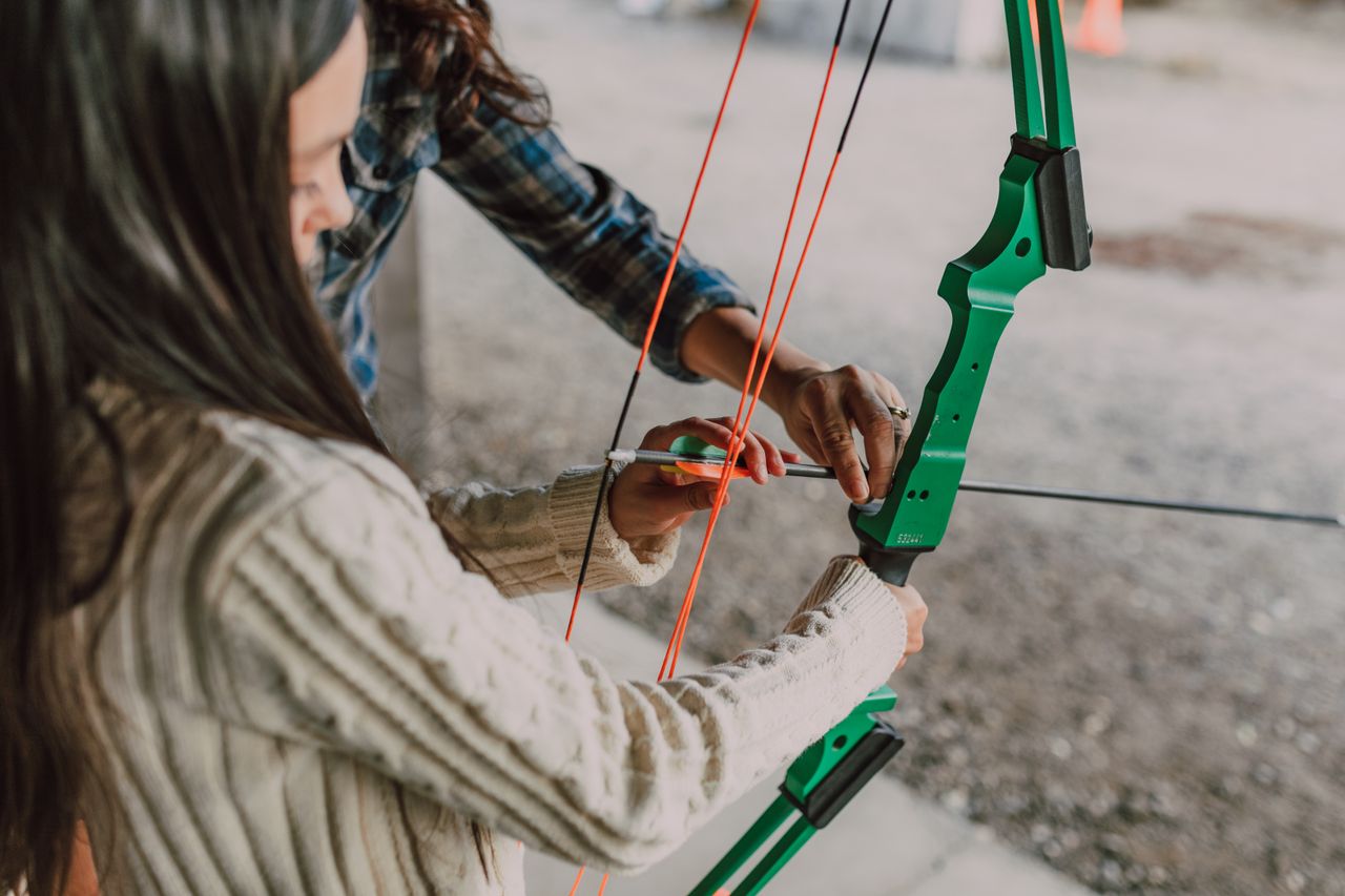 A young girl learning archery with a green recurve bow outdoors, assisted by an adult.