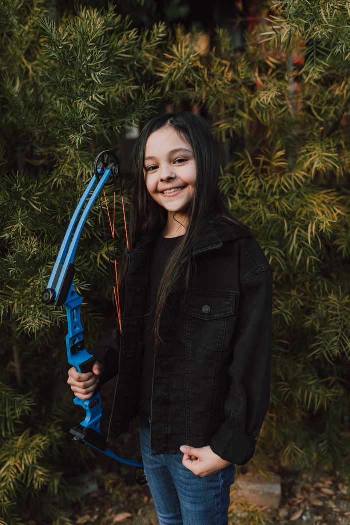 Smiling girl with a blue bow outdoors surrounded by lush greenery, enjoying archery practice.